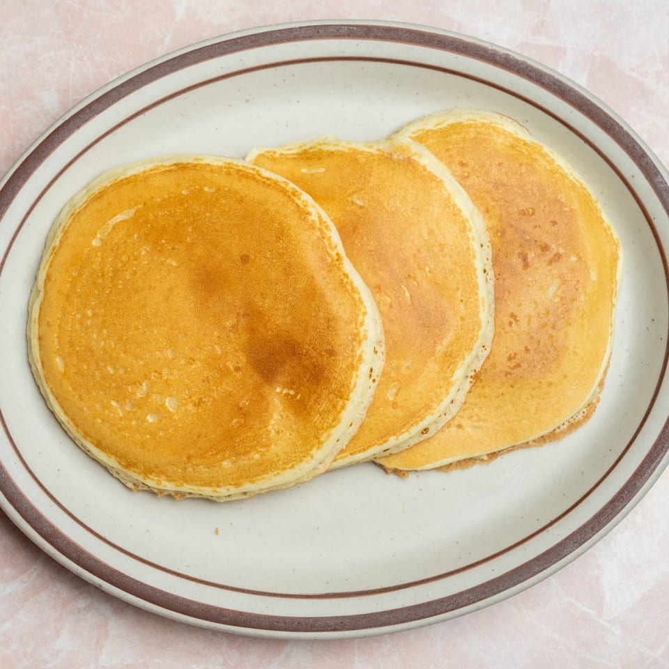 Three golden-brown pancakes overlap on a large vintage plate atop a pale pink surface.