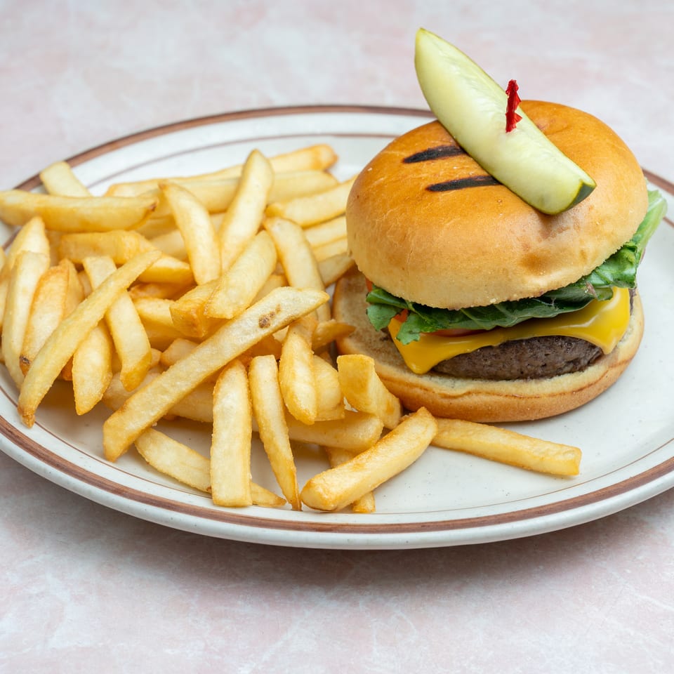 Cheeseburger with lettuce, tomato, and grilled bun, topped with a pickle spear, served alongside golden French fries.