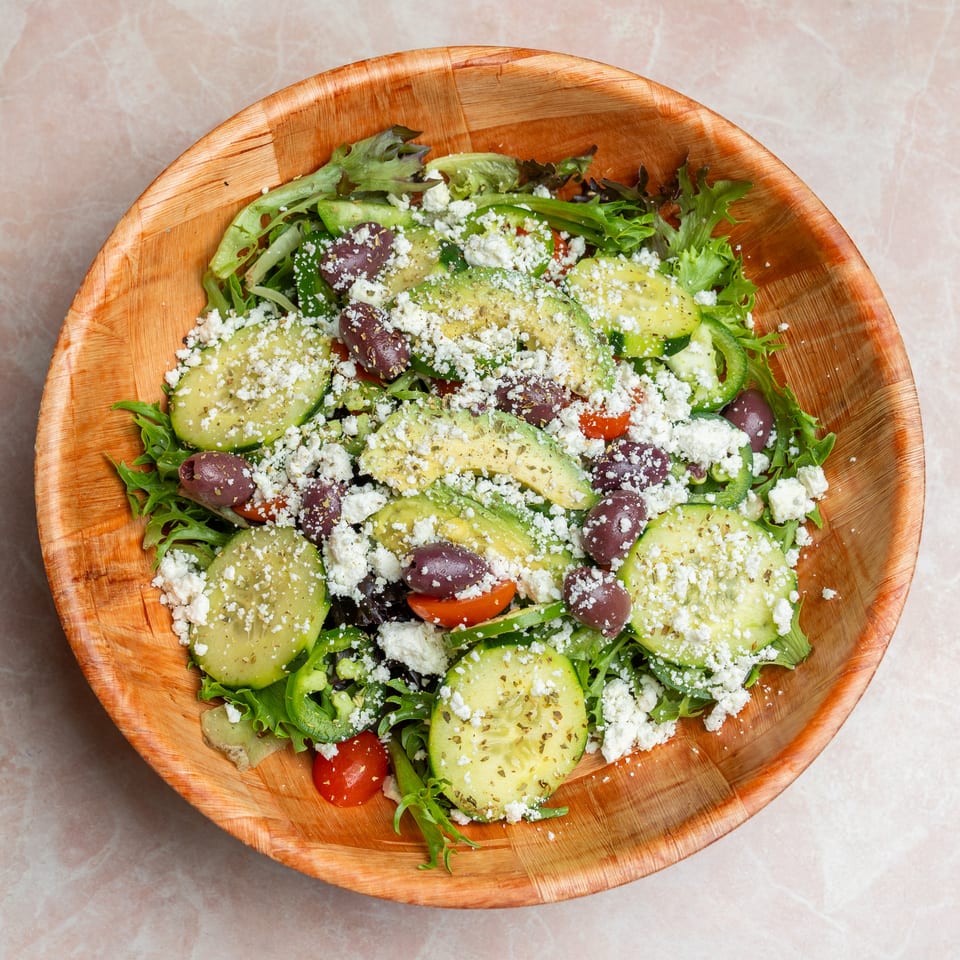 Greek salad with leafy greens, cucumber, tomatoes, olives, avocado, and crumbled feta served in a wooden bowl.