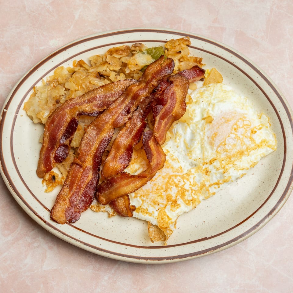 Plate of crispy bacon, fried eggs, and golden home fries on a vintage oval dish.