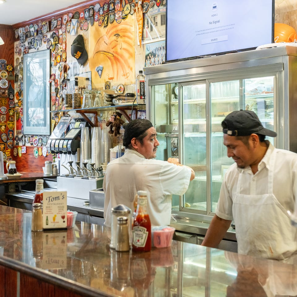 Two restaurant workers stand behind a counter surrounded by colorful patches, a refrigerated display case, condiments, and memorabilia.