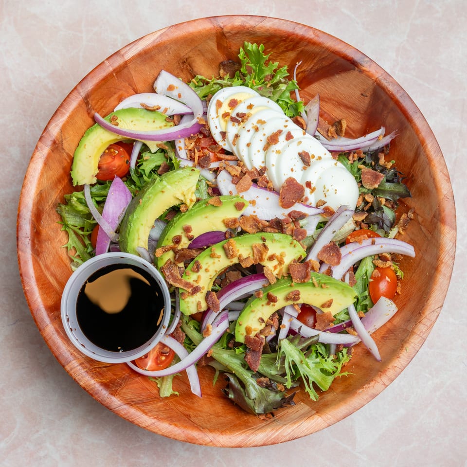 Overhead view of a wooden bowl filled with mixed greens, avocado, hard-boiled eggs, tomatoes, red onion, bacon, and dipping sauce.
