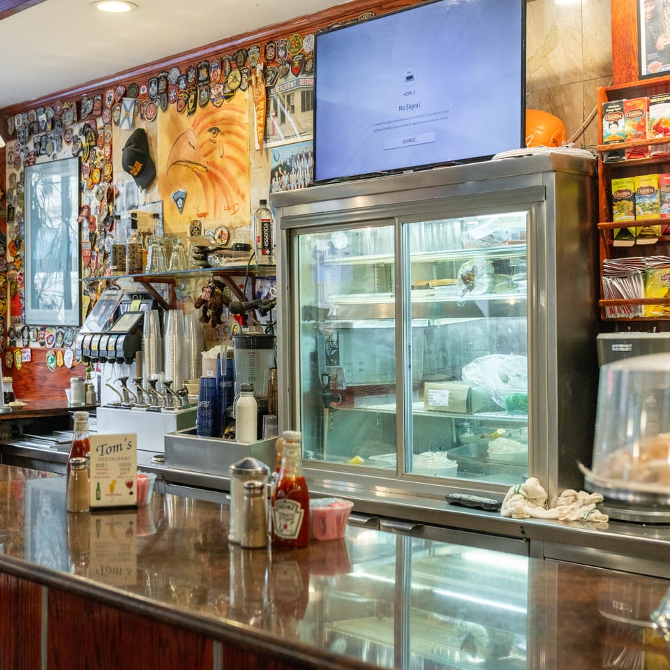 Classic diner counter with a glass food display, soda fountain, tea shelves, condiment bottles, and walls decorated with police badges.