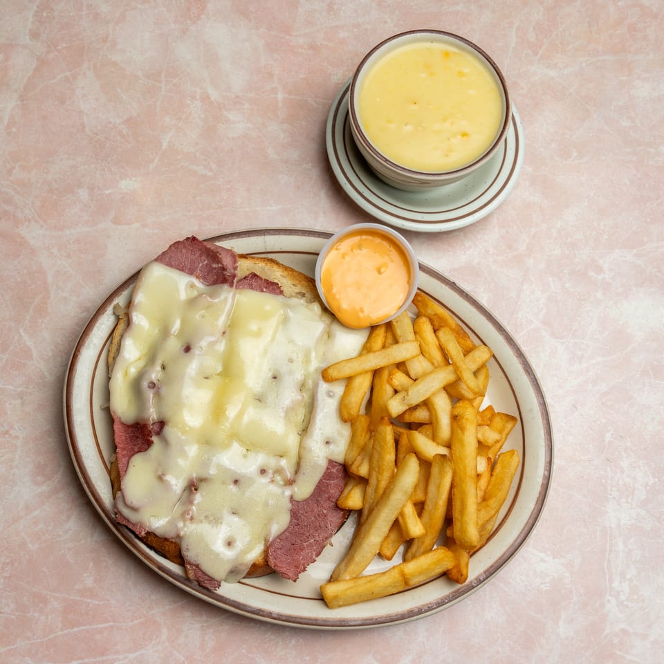 Open-faced corned beef sandwich topped with melted cheese, fries, dipping sauce, and a bowl of creamy soup on a pink tabletop.