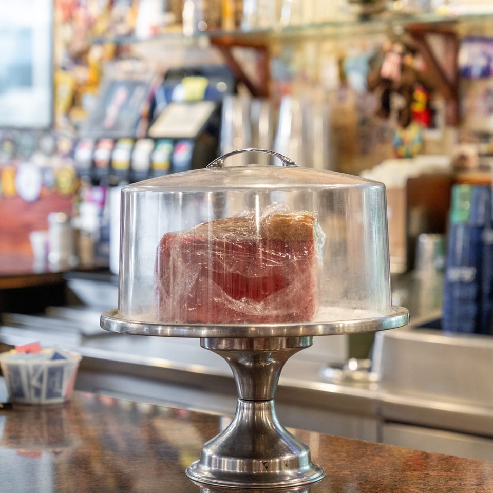 A covered cake stand displays a partially sliced red-and-brown cake on a restaurant counter.