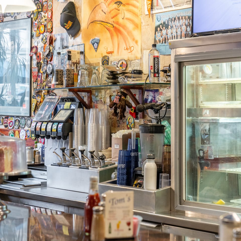 Eclectic restaurant counter with soda fountain, stacked cups, drink dispensers, refrigerated display case, and walls covered in police patches and memorabilia.