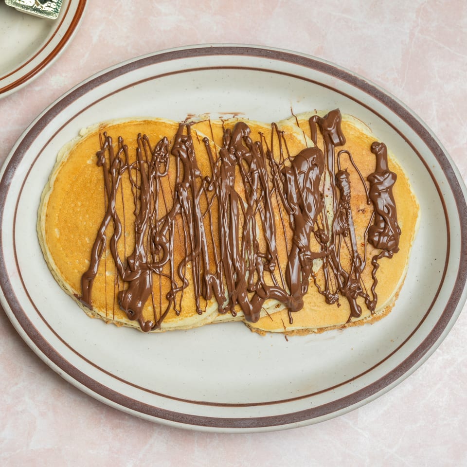 Golden pancakes drizzled with melted chocolate on a large oval plate atop a pink tablecloth.
