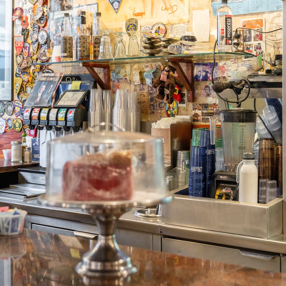 Eclectic restaurant counter with soda dispensers, stacked cups, coffee equipment, memorabilia, currency, and a covered dessert display.