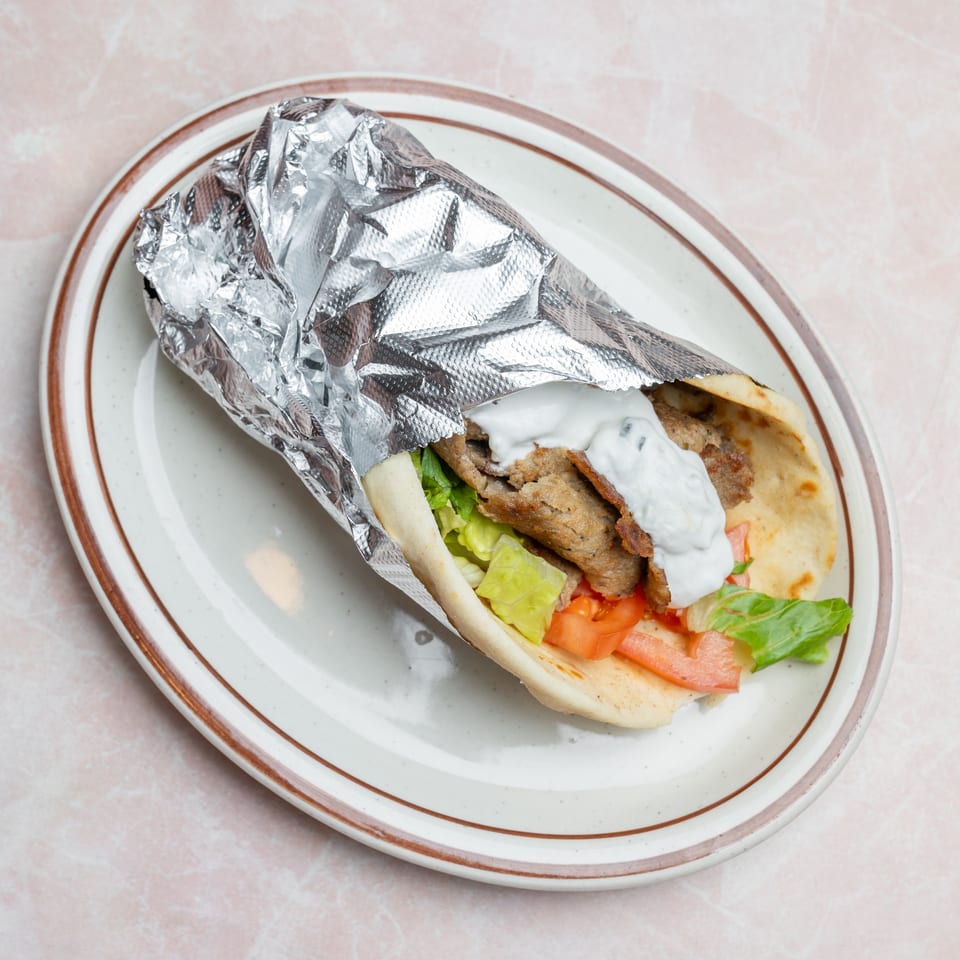 Gyro pita wrapped in foil, filled with sliced meat, lettuce, tomatoes, and white sauce on a ceramic plate.
