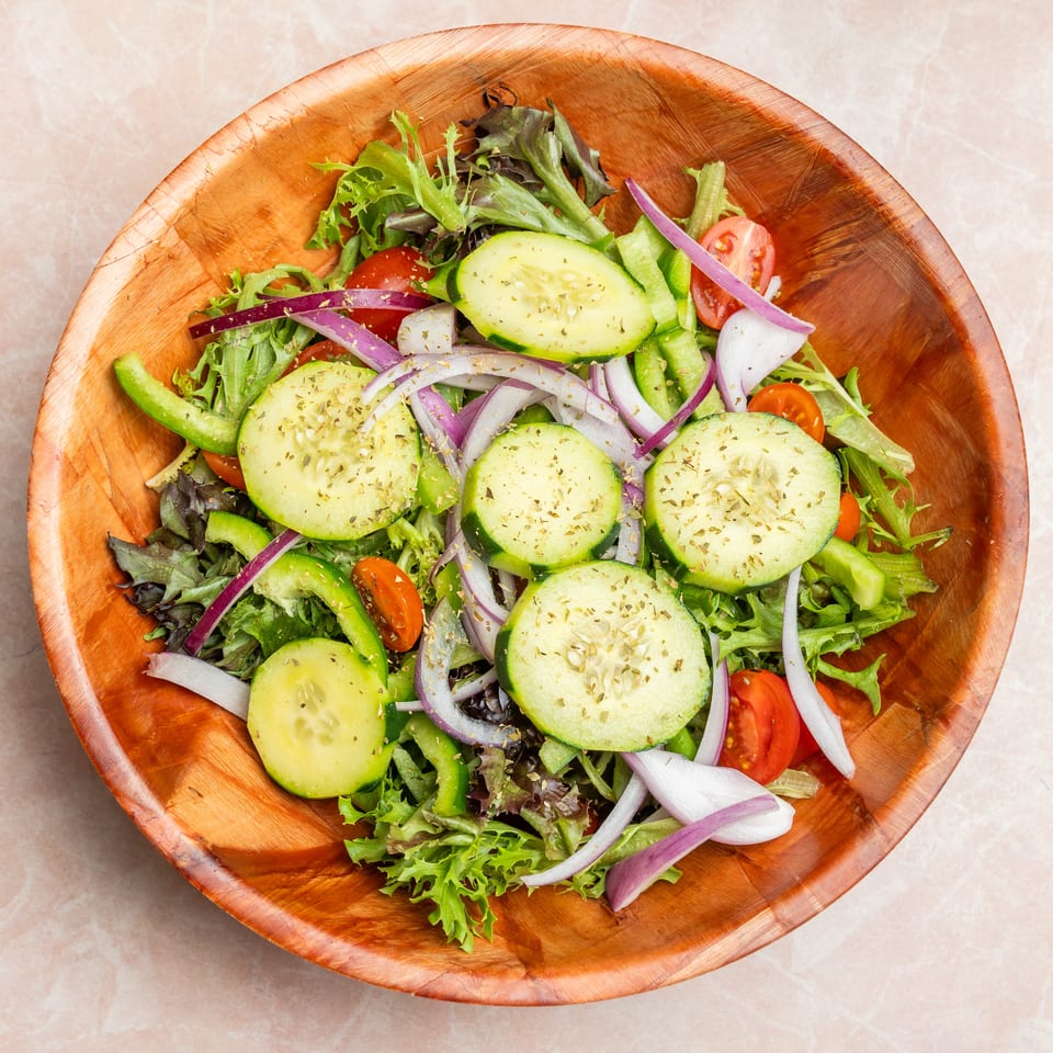 Overhead view of a fresh garden salad with mixed greens, cucumber, tomatoes, green peppers, red onion, and herbs in a wooden bowl.
