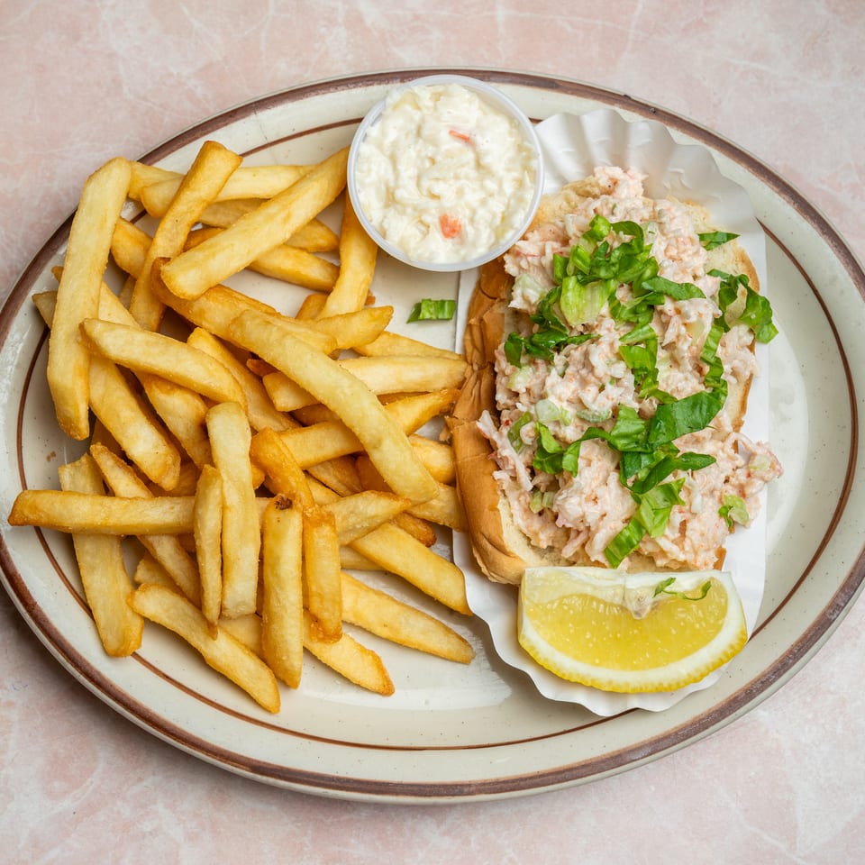 Plate of golden fries with a lobster salad sandwich, coleslaw, fresh herbs, and a lemon wedge.